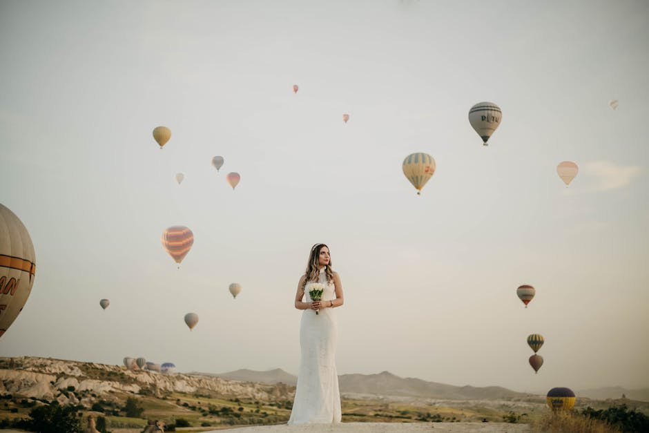 Bride in a white dress standing amidst colorful hot air balloons on a clear day.